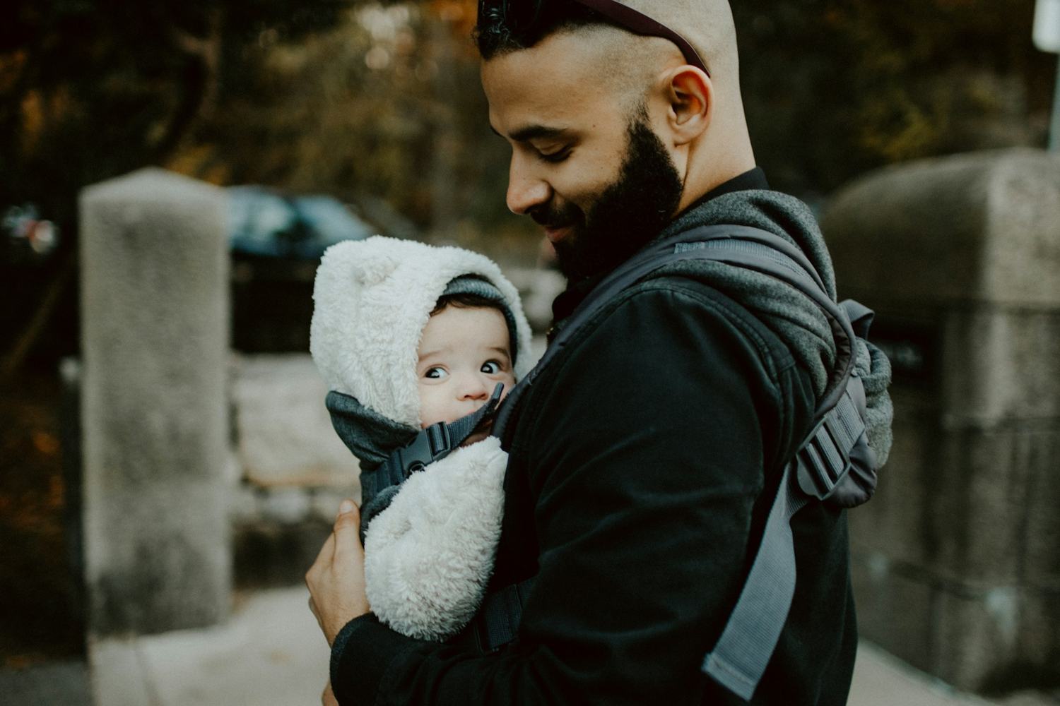 Een man met baard draagt zijn baby in een draagzak door de stad.