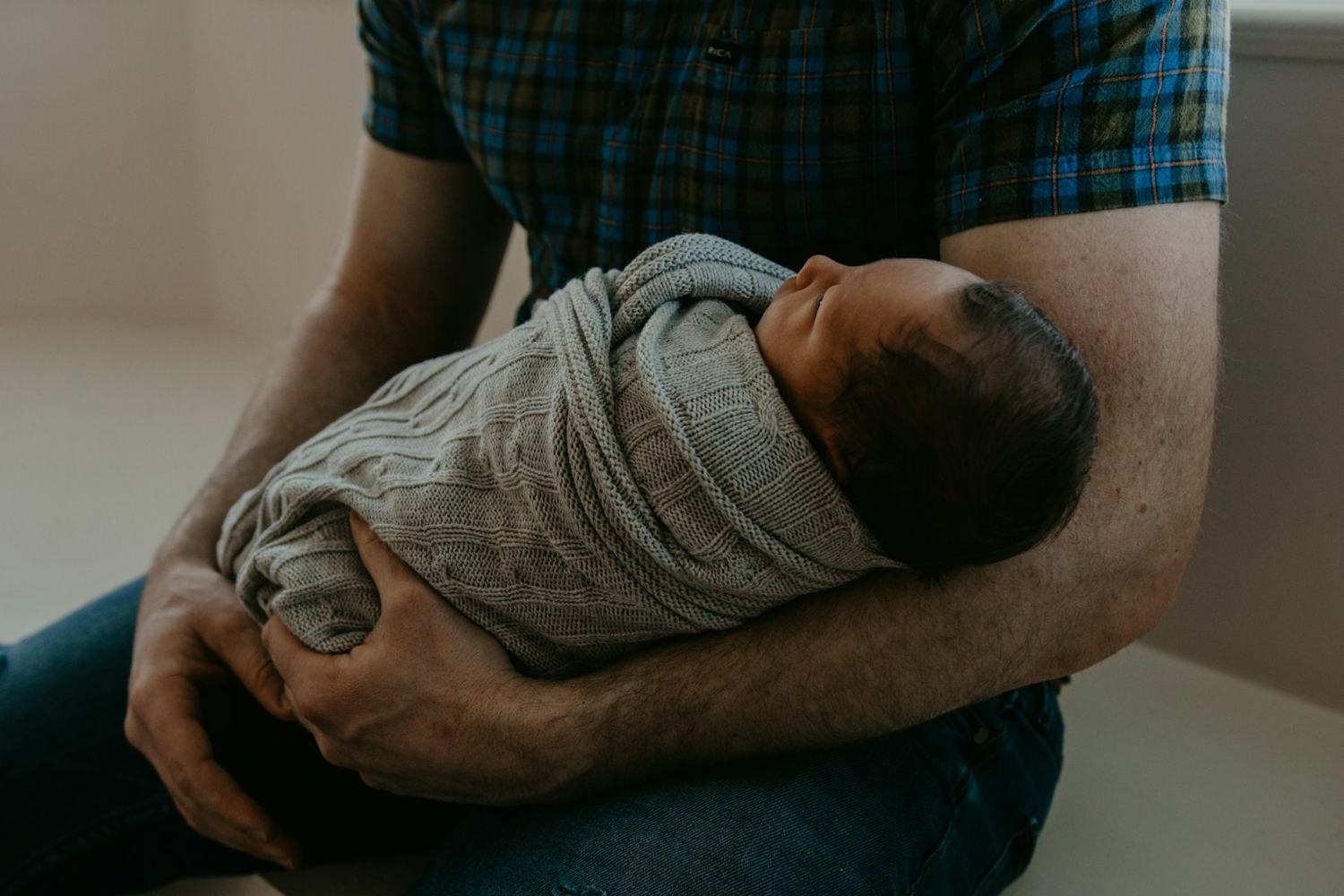 Een man in een geruit overhemd houdt zijn ingebakerde pasgeboren baby vast.